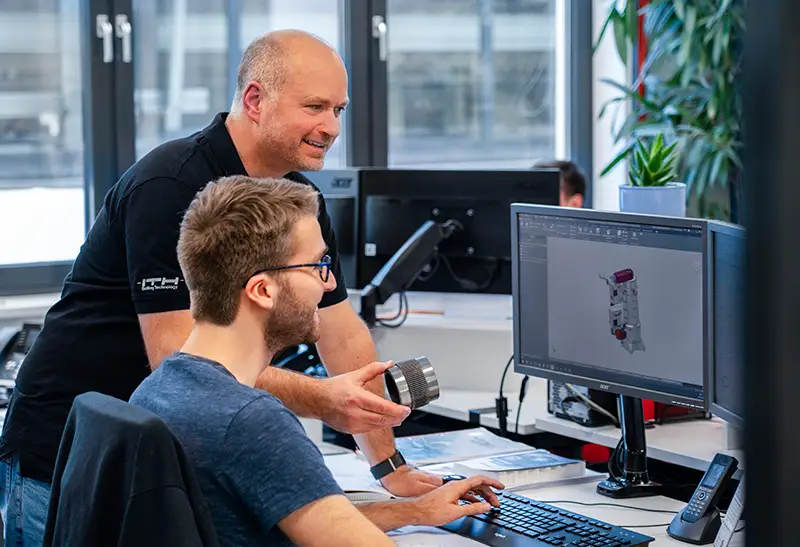 Der Ausbilder erklärt dem Auszubildenden zum technischen Produkdesigner am Schreibtisch ein Bauteil, welches auch im CAD Programm auf dem PC zu sehen ist